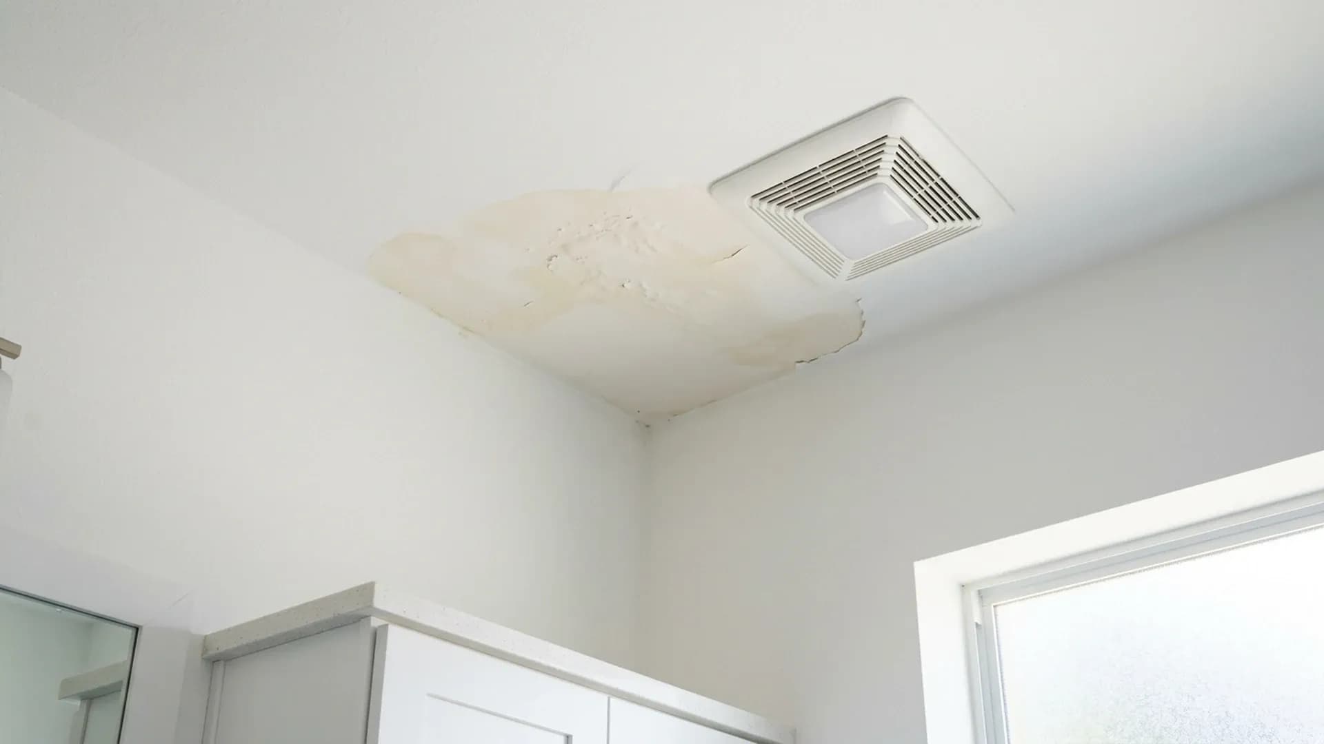 Stained, blistered paint patch at a bathroom ceiling corner beside an exhaust fan grille